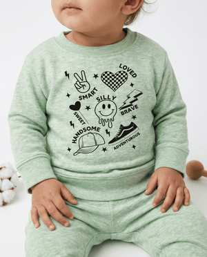A baby wearing a light green heathered long-sleeve sweatshirt and matching sweatpants set, featuring a black graphic print with words like 'LOVED', 'SMART', 'SILLY', 'BRAVE', and 'HANDSOME' surrounding playful icons.