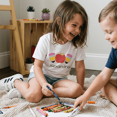 A young girl sitting on a beige knit blanket in a living room, wearing a white personalized graphic T-shirt with a colorful apple and a custom name printed on the front. She is laughing and holding a blue crayon, interacting with a boy who is partially visible on the right.
