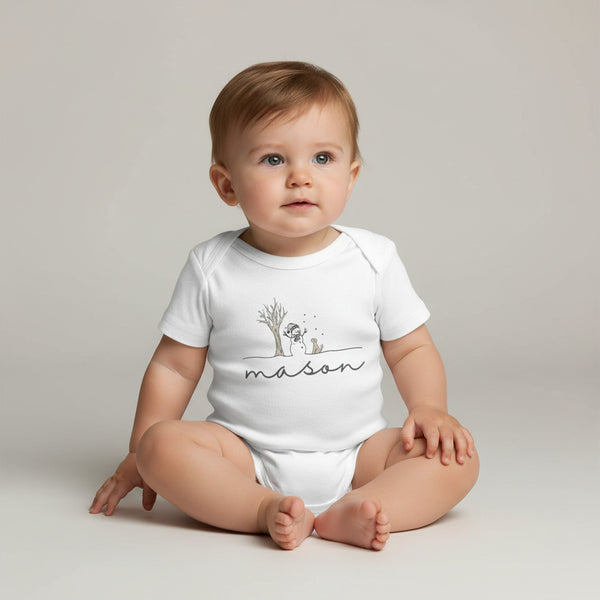 A baby sitting cross-legged wearing a white short-sleeve bodysuit with a minimalist tree graphic and the name 'mason' printed across the chest, photographed against a plain light gray studio background.