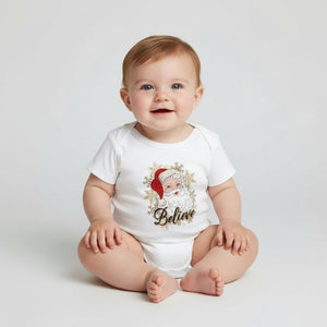 A baby sitting on a light gray studio floor wearing a white short-sleeve bodysuit with a festive Santa graphic and the word 'Believe' printed on the front.