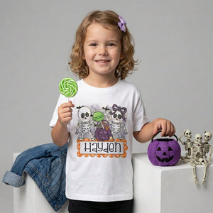 A young girl with curly blonde hair wearing a white graphic T-shirt with Cute Halloween skeletons and candy design, with a custom name on it. 