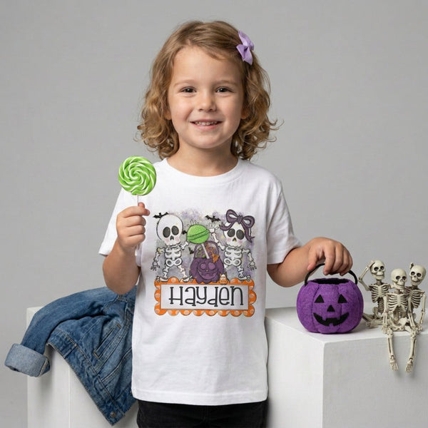 A young girl with curly blonde hair wearing a white graphic T-shirt with Cute Halloween skeletons and candy design, with a custom name on it. 