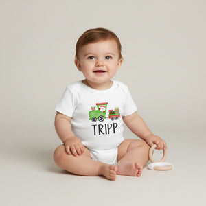 A baby sitting on a light beige studio floor wearing a white short-sleeve bodysuit with a colorful cartoon train graphic and the name 'TRIPP' printed below it. The baby is holding a light wood teething ring.