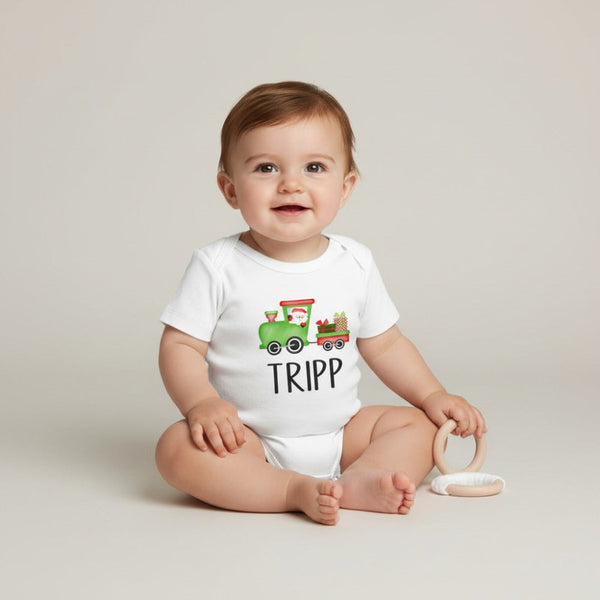 A baby sitting on a light beige studio floor wearing a white short-sleeve bodysuit with a colorful cartoon train graphic and the name 'TRIPP' printed below it. The baby is holding a light wood teething ring.