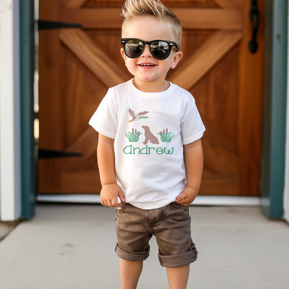 A young boy wearing a white graphic t-shirt with a duck hunting design and the name 'Andrew' in green, paired with brown shorts and black sunglasses, standing in front of a wooden barn door.