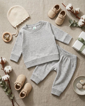 A baby two-piece loungewear set in light heather gray, featuring a long-sleeve crewneck sweatshirt and matching jogger pants, styled in a flat lay with a cream knit hat, beige baby shoes, cotton stems, and a gift box on a beige linen background.
