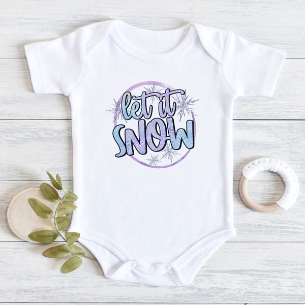 A white short-sleeve baby bodysuit laid flat on a light wood surface, featuring a circular graphic that reads 'let it SNOW' in blue and purple lettering with snowflake details. A small green leaf sprig and a white wooden teething ring are placed beside it as styling props.