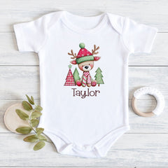 A white short-sleeve baby bodysuit laid flat on a light wood surface, printed with a cute holiday reindeer wearing a red and green hat and scarf, with the name 'Taylor' below. Decorative leaves and a wooden teether are placed beside it.
