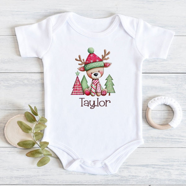 A white short-sleeve baby bodysuit laid flat on a light wood surface, printed with a cute holiday reindeer wearing a red and green hat and scarf, with the name 'Taylor' below. Decorative leaves and a wooden teether are placed beside it.