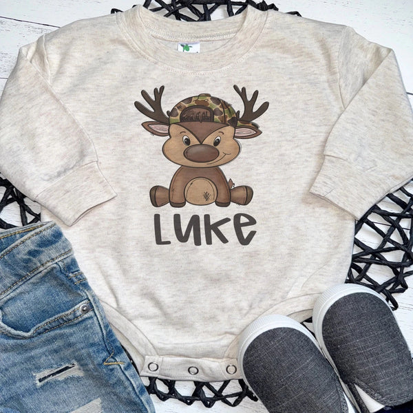 A baby long-sleeve onesie in a light oatmeal heather fabric, featuring a cartoon reindeer wearing a camouflage hat and the name 'LUKE' printed below. The onesie is styled in a flat lay with blue jeans and gray slip-on shoes on a black woven mat over a white wood background.