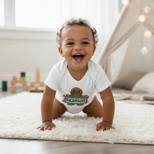 A smiling baby crawling on a white rug in a bright nursery, wearing a white short-sleeve bodysuit with a gingerbread cookie graphic and the name 'Grayson' printed on the front.
