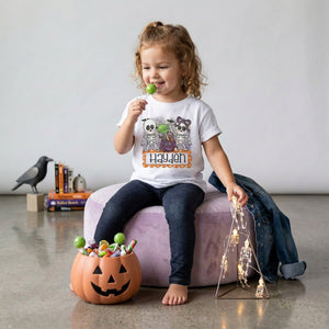 A young girl sits on a lavender ottoman in a styled room, wearing a white graphic t-shirt with cartoon ghosts and the name 'Hayden' in orange. She is holding a green lollipop, with a jack-o'-lantern candy bucket, books, a crow figurine, and string lights in the background.