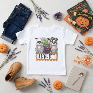 A white kids' short-sleeve T-shirt with a Halloween graphic of two cartoon skeletons and the name 'Hayden' in orange, styled in a flat lay with jeans, shoes, pumpkins, lavender, candy, and books on a light surface.