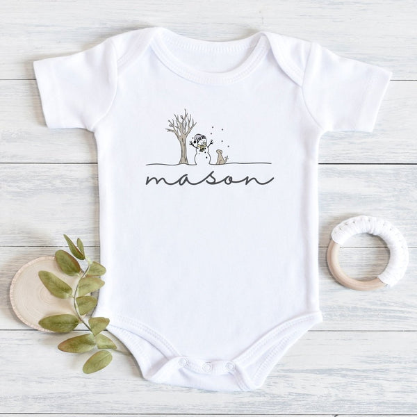 A white short-sleeve baby bodysuit laid flat on a light wood surface, printed with a simple winter scene and a custom name in black script. A small leafy branch and a white teething ring are placed beside it as styling props.