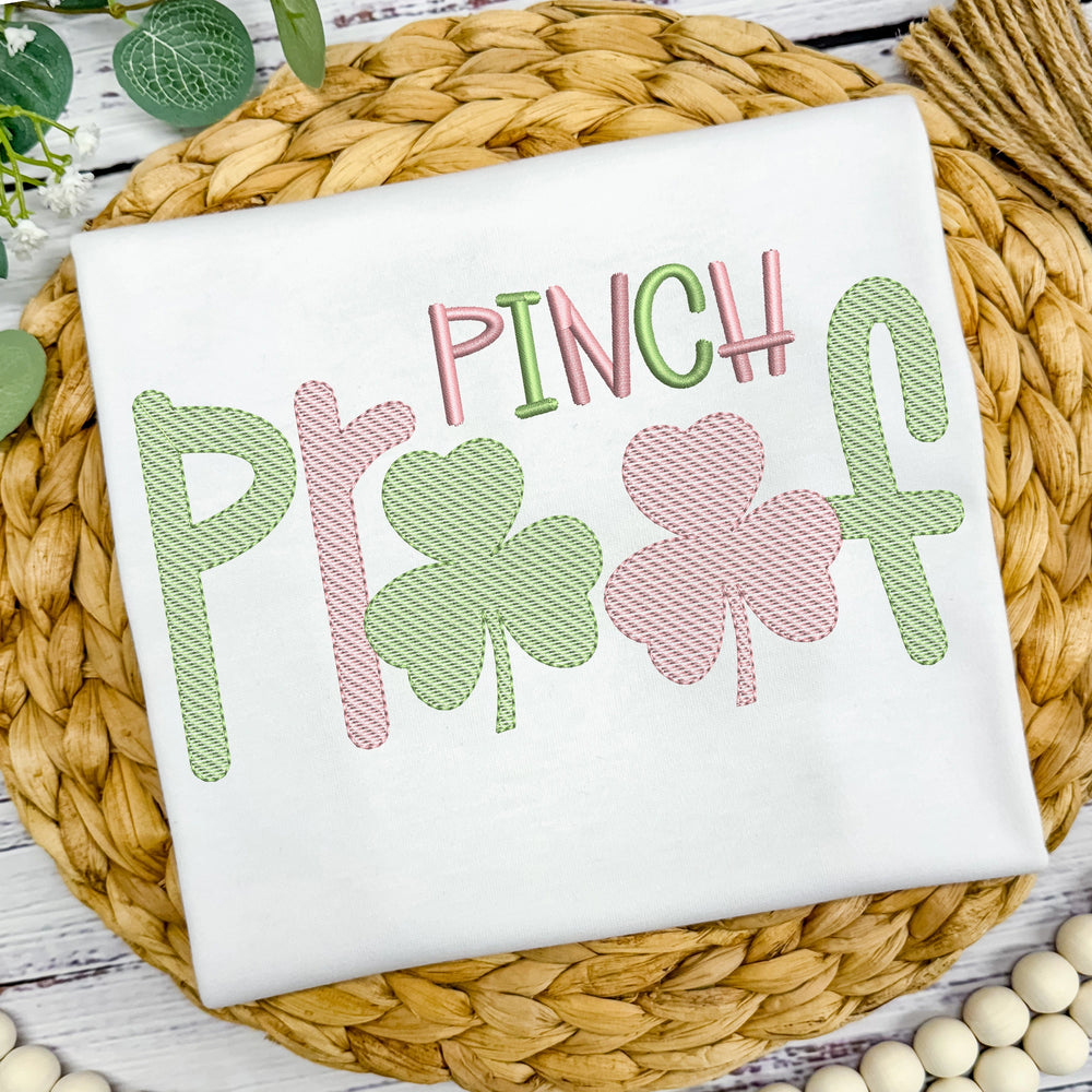White t-shirt with 'Pinch Play' text and shamrock designs on a woven mat.
