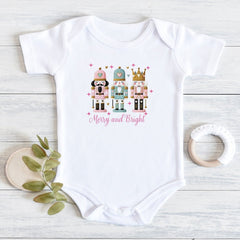 A white short-sleeve baby bodysuit laid flat on a light wood surface, featuring a colorful nutcracker illustration and the words 'Merry and Bright' on the chest. A small green leafy sprig and a white teething ring are placed beside it as styling props.