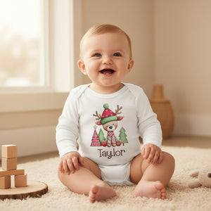 A smiling baby sits on a cream carpet wearing a white long-sleeve bodysuit with a festive Christmas deer graphic and the name 'Taylor' printed below. The scene is set in a warm, softly lit room with wooden toys and a vase in the background.