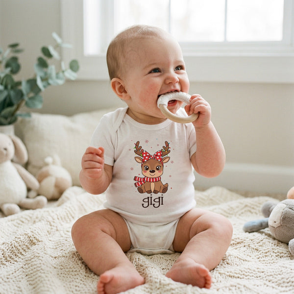A baby sitting on a cream knit blanket in a bright room, wearing a white bodysuit with a cartoon reindeer graphic and the name 'jigi' printed below it.