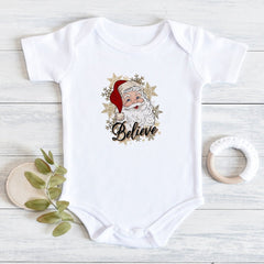 A white short-sleeve baby bodysuit laid flat on a light wood surface, featuring a festive Santa Claus graphic with the word 'Believe' in script. A wooden teething ring and green leaves are placed beside it as styling props.