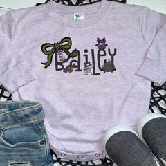 A light lavender heather short-sleeve baby bodysuit with a custom 'Bailey' Halloween graphic, styled in a flat lay with blue jeans and gray sneakers on a black lattice mat.