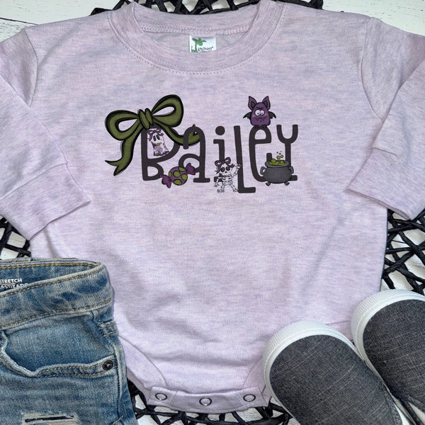 A light lavender heather short-sleeve baby bodysuit with a custom 'Bailey' Halloween graphic, styled in a flat lay with blue jeans and gray sneakers on a black lattice mat.