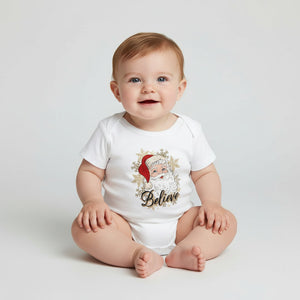 A baby sitting on a light gray studio floor wearing a white short-sleeve bodysuit with a festive Santa graphic and the word 'Believe' printed on the front.