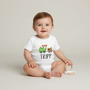 A baby sitting on a light beige studio floor wearing a white short-sleeve bodysuit with a colorful cartoon train graphic and the name 'TRIPP' printed below it. The baby is holding a light wood teething ring.