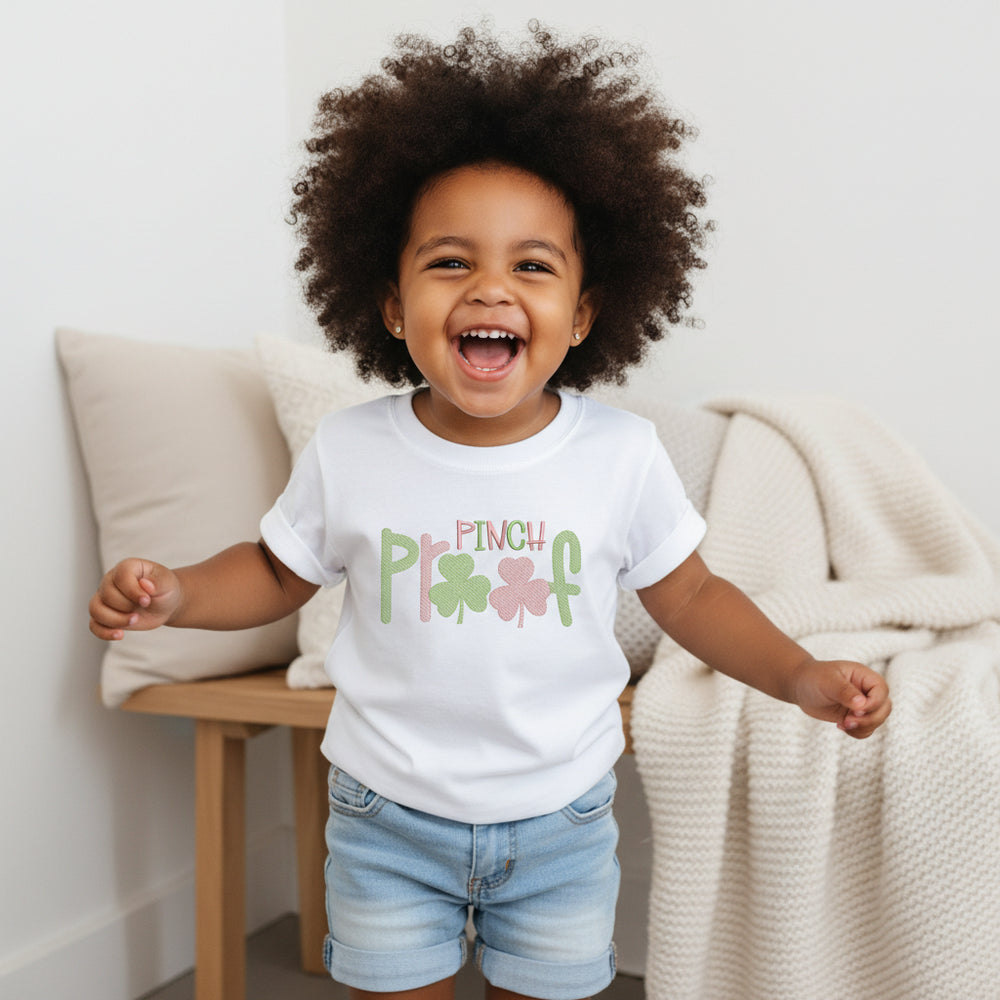 White t-shirt with 'Pinch Play' text and shamrock designs on a woven mat.