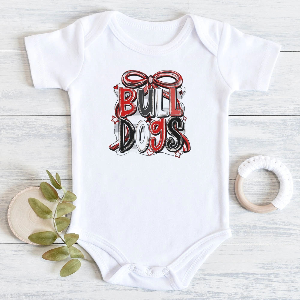 A white short-sleeve baby bodysuit laid flat on a light wood surface, featuring a red, black, and white 'BULL DOGS' graphic with a bow on the chest. A small green leaf sprig, a wooden coaster, and a wooden teething ring are placed around it as styling props.