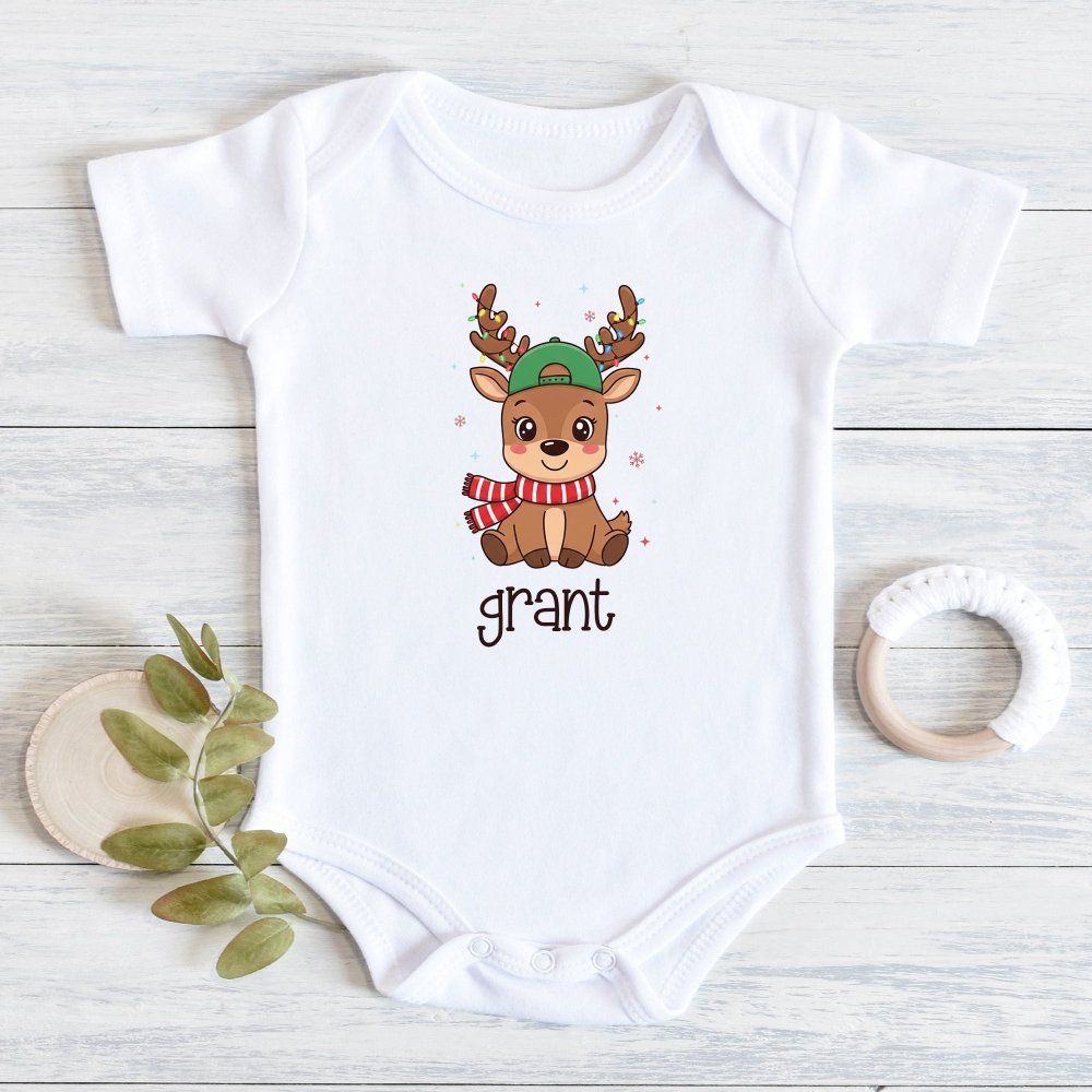 A white short-sleeve baby bodysuit laid flat on a light wood surface, featuring a cartoon reindeer wearing a green hat and red-and-white striped scarf, with the name 'grant' printed below. A small leafy sprig and a white teething ring are placed beside it as styling props.