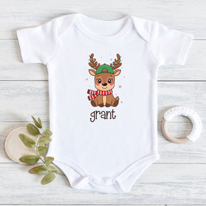 A white short-sleeve baby bodysuit laid flat on a light wood surface, featuring a cartoon reindeer wearing a green hat and red-and-white striped scarf, with the name 'grant' printed below. A small leafy sprig and a white teething ring are placed beside it as styling props.