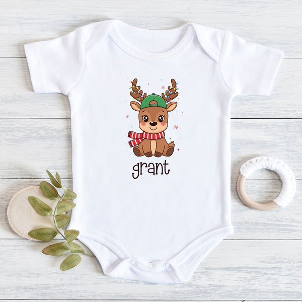 A white short-sleeve baby bodysuit laid flat on a light wood surface, featuring a cartoon reindeer wearing a green hat and red-and-white striped scarf, with the name 'grant' printed below. A small leafy sprig and a white teething ring are placed beside it as styling props.