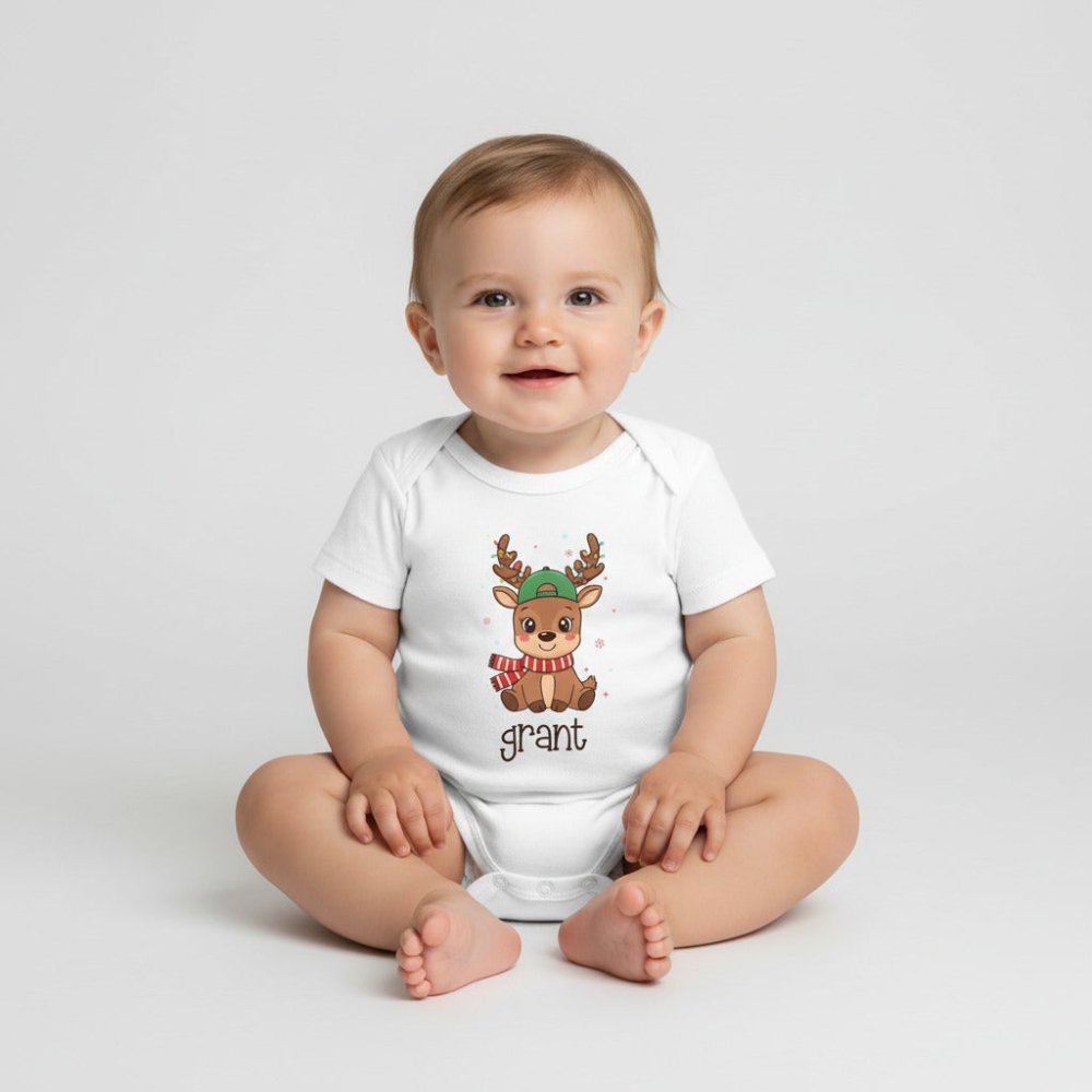 A white short-sleeve baby bodysuit laid flat on a light wood surface, featuring a cartoon reindeer wearing a green hat and red-and-white striped scarf, with the name 'grant' printed below. A small leafy sprig and a white teething ring are placed beside it as styling props.