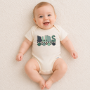 Baby wearing a onesie with 'BUBS' printed on it, sitting on a soft surface.