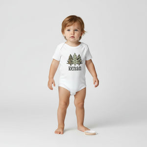 A toddler standing barefoot on a light gray studio background, wearing a white short-sleeve bodysuit with a pine tree graphic and the name 'kenan' printed on the front.