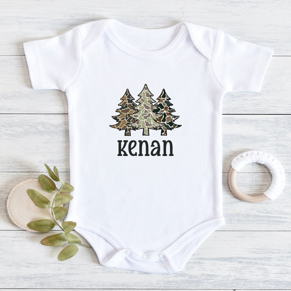 A white short-sleeve baby bodysuit lay flat on a light wood surface, printed with three camouflage-patterned pine trees and a custom name printed on it. A small green leafy sprig and a wooden teething ring are placed beside it as props.