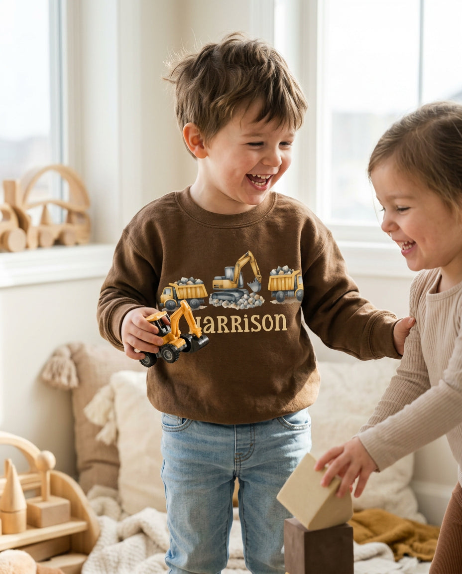 A young boy wearing a brown crewneck sweatshirt with a construction graphic and the name 'HARRISON' printed on the front, standing in a bright room with a toddler next to him.