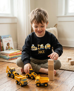 A young child sitting on a wooden floor in a bright room, wearing a black crewneck sweatshirt with a construction-themed graphic and the name 'JACE' printed on the front. The child is playing with yellow toy trucks and wooden blocks.