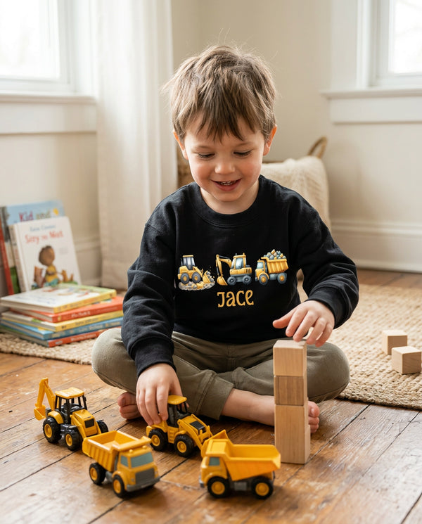 A young child sitting on a wooden floor in a bright room, wearing a black crewneck sweatshirt with a construction-themed graphic and the name 'JACE' printed on the front. The child is playing with yellow toy trucks and wooden blocks.