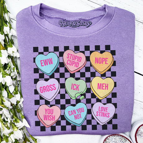 Folded purple sweatshirt with colorful heart-shaped sayings on a white surface with flowers.