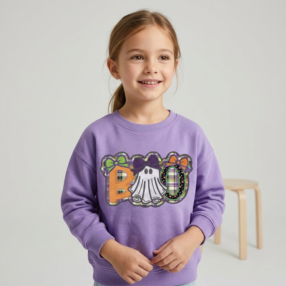 A young girl with light brown hair in a ponytail smiles while wearing a lavender crewneck sweatshirt. The sweatshirt features a colorful 'RO' graphic with a ghost, plaid patterns, and bows.