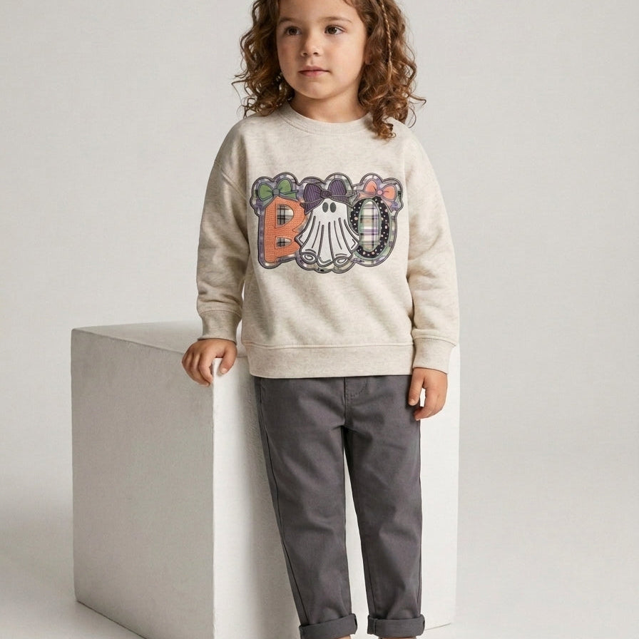 A young child stands on a white pedestal wearing a cream crewneck sweatshirt with a colorful embroidered 'BOO' design, dark gray pants, and white slip-on shoes against a light gray studio background.