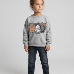 A young child stands against a light gray studio background wearing a heather gray sweatshirt with a colorful 'BOO' graphic and dark blue jeans.