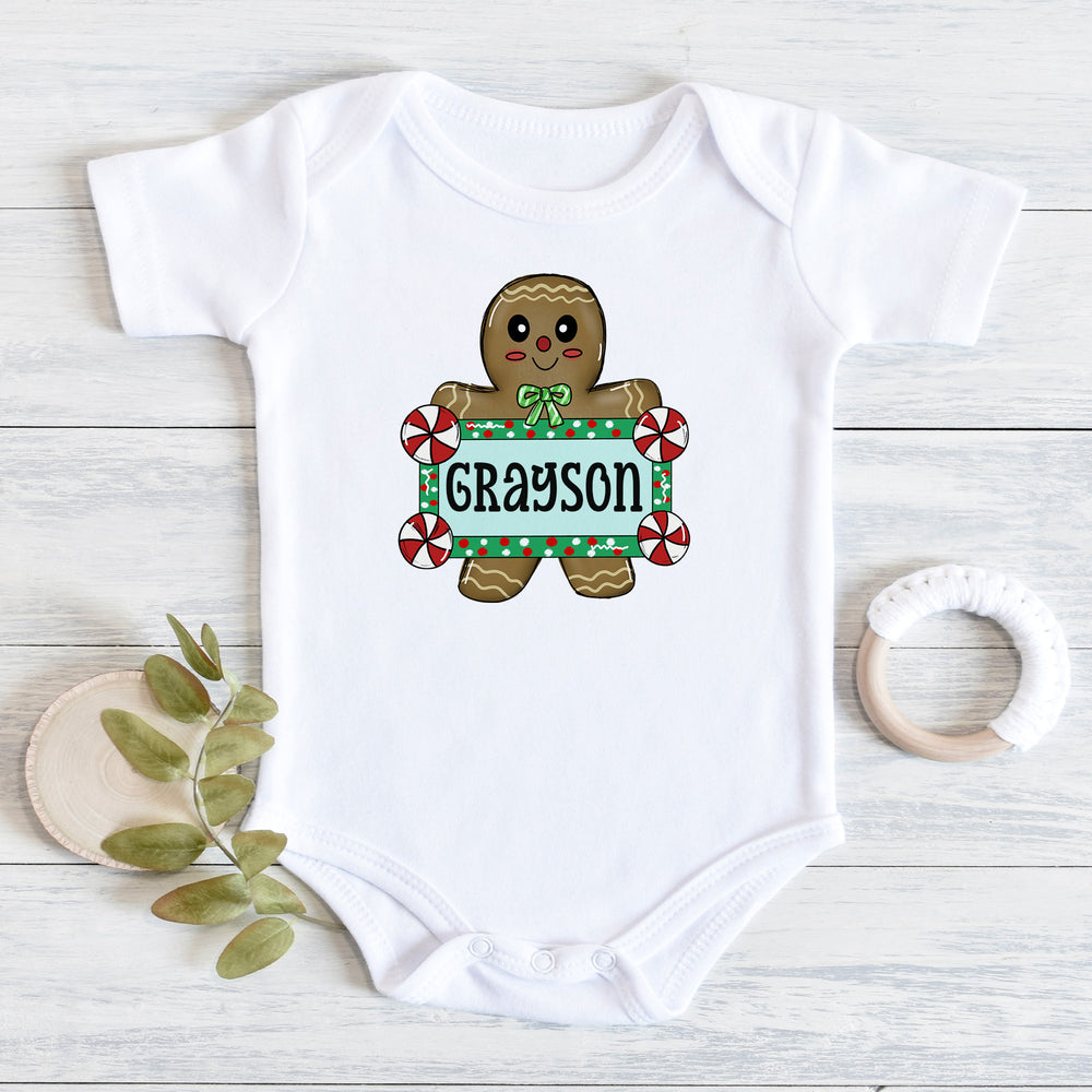 A white short-sleeve baby bodysuit laid flat on a light wood surface, featuring a gingerbread man graphic holding a personalized name sign that reads 'Grayson.' Decorative props including a small leafy sprig and a white teething ring are placed around the bodysuit.