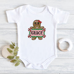 A white short-sleeve baby bodysuit laid flat on a light wood surface, featuring a gingerbread cookie graphic holding a sign that says 'GRACE' in red, green, and white. A small leafy sprig and a white teething ring are placed beside the bodysuit as styling props.