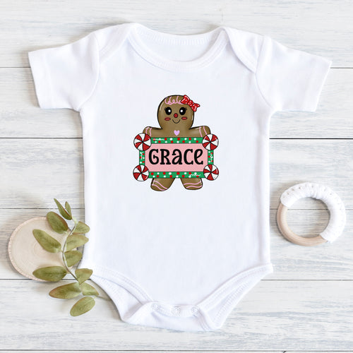 A white short-sleeve baby bodysuit laid flat on a light wood surface, featuring a gingerbread cookie graphic holding a sign that says 'GRACE' in red, green, and white. A small leafy sprig and a white teething ring are placed beside the bodysuit as styling props.