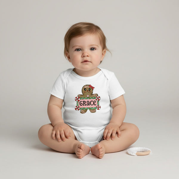 A baby sitting cross-legged on a light gray studio background, wearing a white short-sleeve bodysuit with a gingerbread man graphic holding a sign that says 'GRACE'.