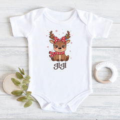 A white short-sleeve baby bodysuit laid flat on a light wood surface, featuring a cute cartoon deer graphic with a red bow and striped scarf above the name 'Jigi'. A small wooden leaf holder and a white teething ring are placed beside it as props.