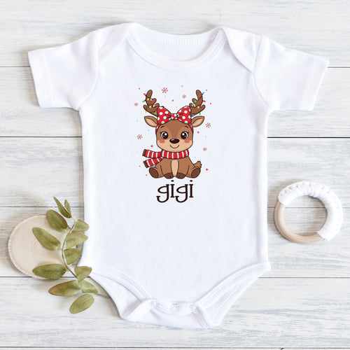 A white short-sleeve baby bodysuit laid flat on a light wood surface, featuring a cute cartoon deer graphic with a red bow and striped scarf above the name 'Jigi'. A small wooden leaf holder and a white teething ring are placed beside it as props.