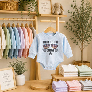 A light blue long-sleeve baby bodysuit with a 'TALK TO ME GOOSE' graphic print hanging on a wooden hanger. The bodysuit is displayed on a wooden clothing rack in a styled nursery-like setting with other colorful baby clothes, folded shirts, and potted plants.